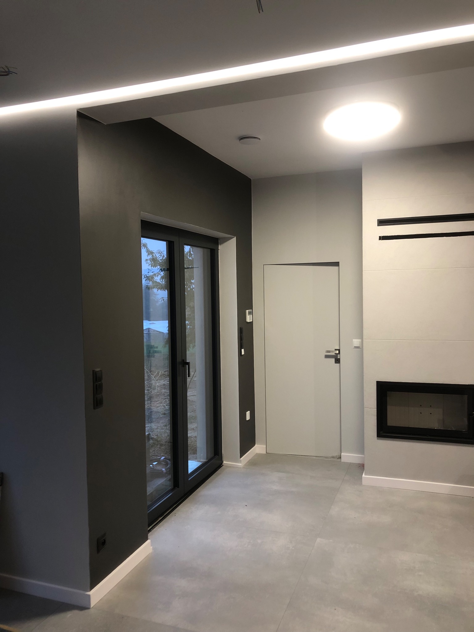 Nowoczesne wnętrze z szarą ścianą, przesuwnymi drzwiami balkonowymi, minimalistycznym kominkiem i oświetleniem LED w suficie.