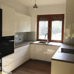 Nowoczesna kuchnia z białymi, lakierowanymi frontami szafek, drewnianym blatem i oknem z drewnianą ramą, widok z perspektywy narożnika, logo firmy Glass Effect w lewym dolnym rogu.