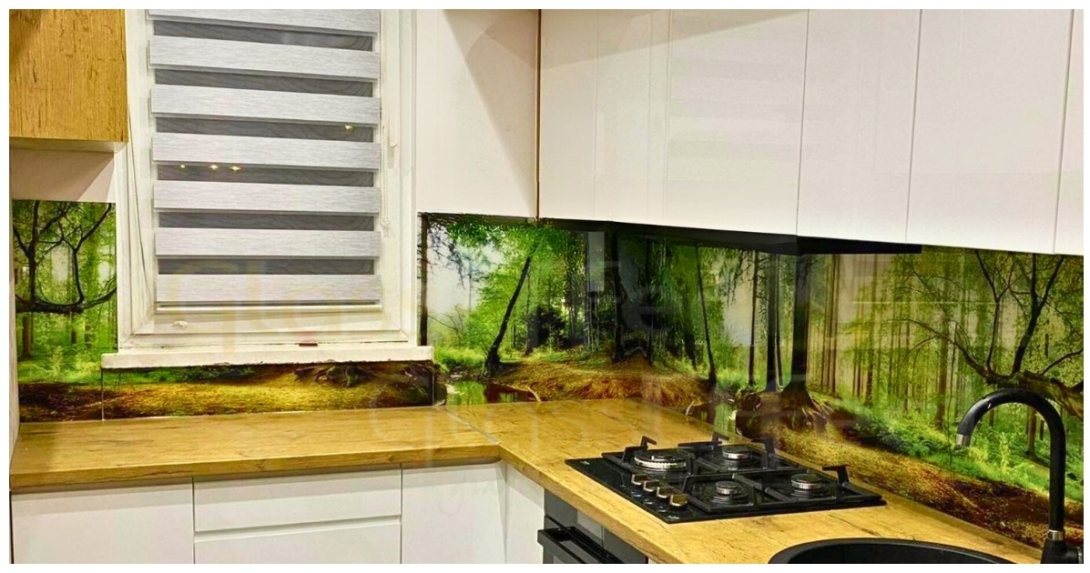 Kuchnia z blatem imitującym drewno i szklanym panelem ściennym z motywem leśnym, widoczne białe szafki i czarna płyta gazowa.