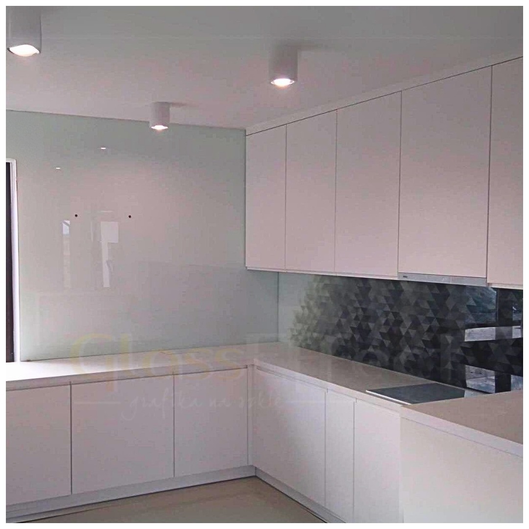 Biała kuchnia z przeszklonym panelem ściennym i szafkami bez uchwytów, oświetlona punktowymi lampami sufitowymi.