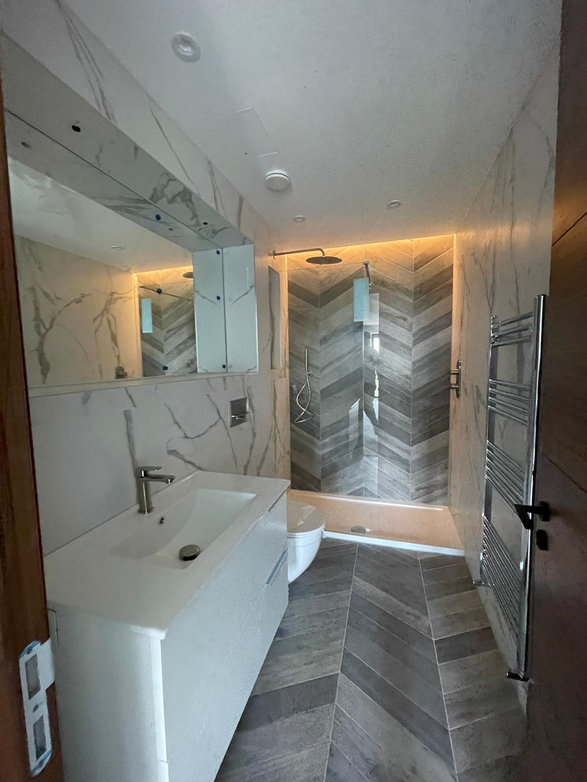 Wykończona łazienka z marmurowymi płytkami, wzór jodełki na podłodze i ścianie prysznica, podświetlenie LED w strefie prysznicowej, biała umywalka z szafką i toaleta.