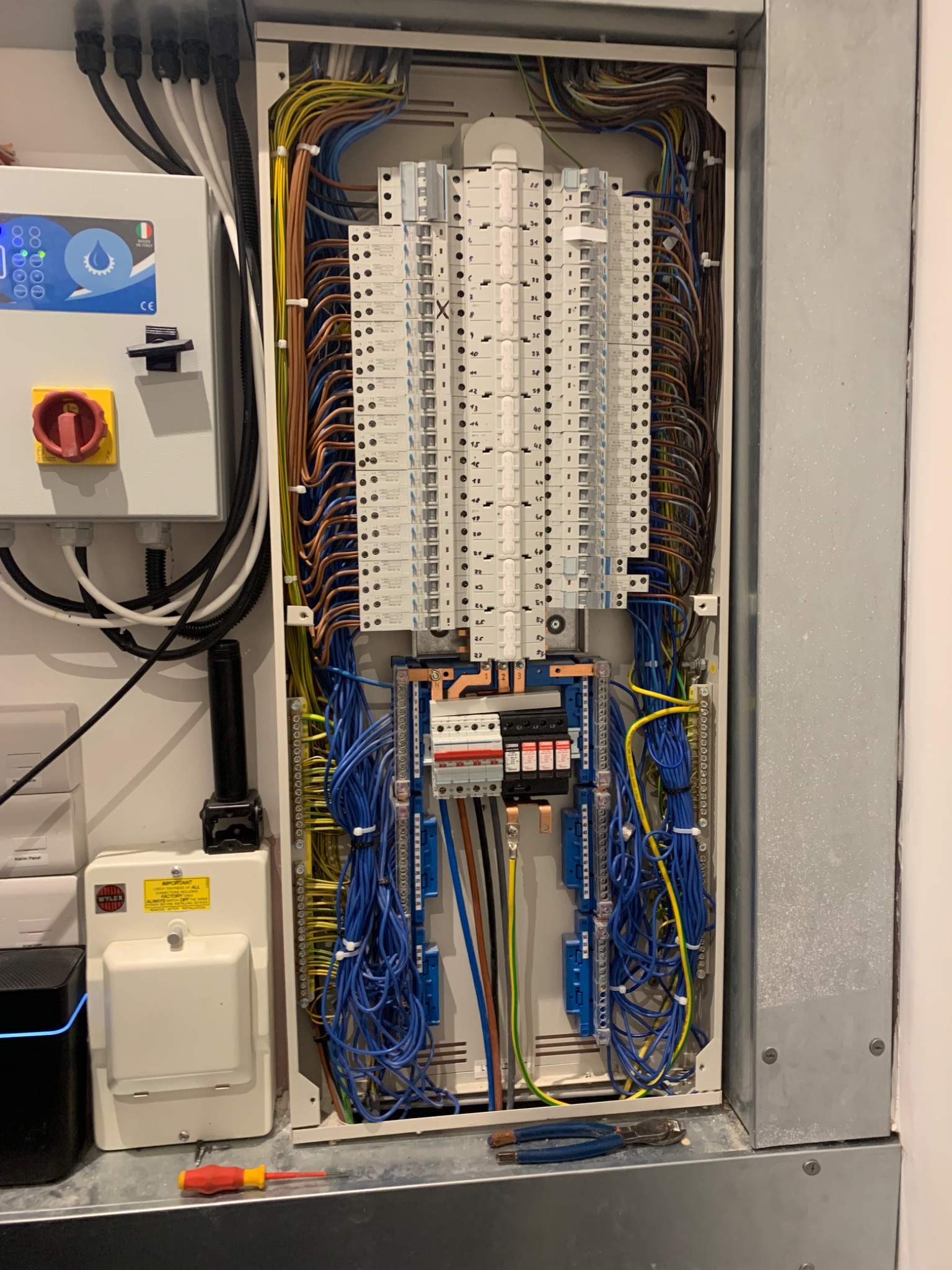 Otwarta skrzynka elektryczna z licznymi przewodami, bezpiecznikami i elementami sterującymi, uzupełniona o narzędzia - śrubokręt i kombinerki.