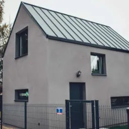 Nowoczesny dom jednorodzinny z szarą elewacją, metalowym dachem i ogrodzeniem panelowym, widziany z zewnątrz w pochmurny dzień. W oknie widoczny neon z napisem 'SOSN'.