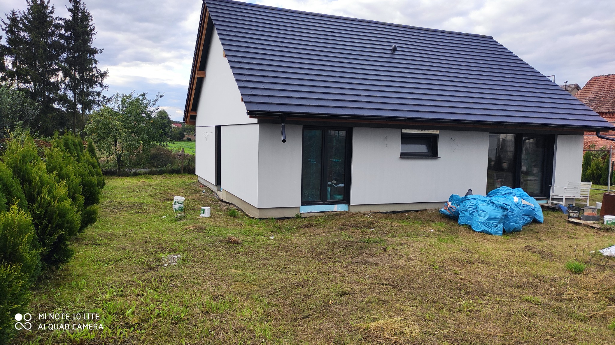 Jednopiętrowy dom z białą elewacją, ciemną dachówką i czarnymi oknami, otoczony trawnikiem, z widocznymi workami z odpadami budowlanymi i pojemnikami po farbie, sugerujący prace wykończeniowe.