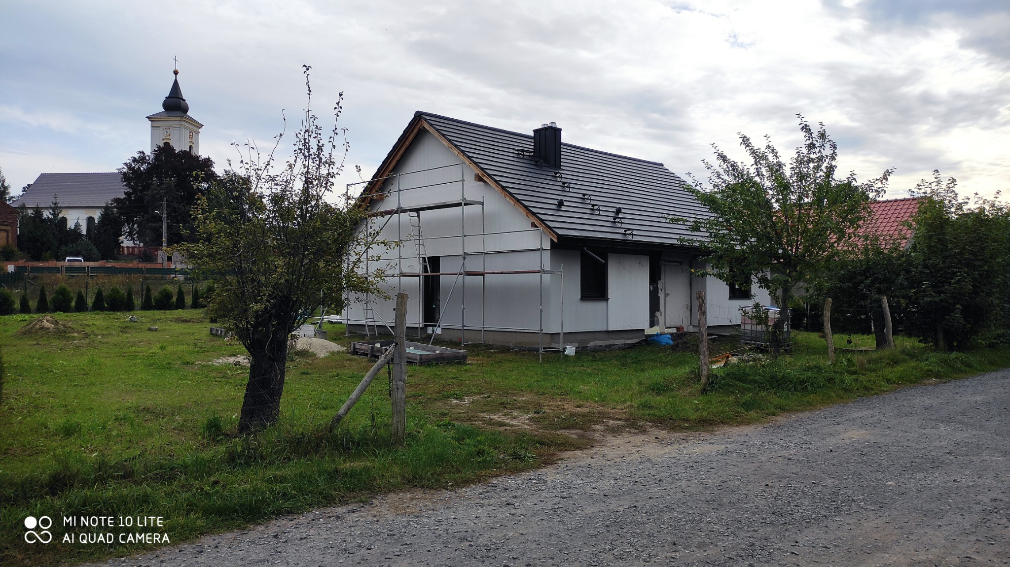 Dom jednorodzinny w trakcie prac elewacyjnych, z widocznym rusztowaniem i częściowo ocieploną elewacją, na tle wiejskiego krajobrazu z kościołem.
