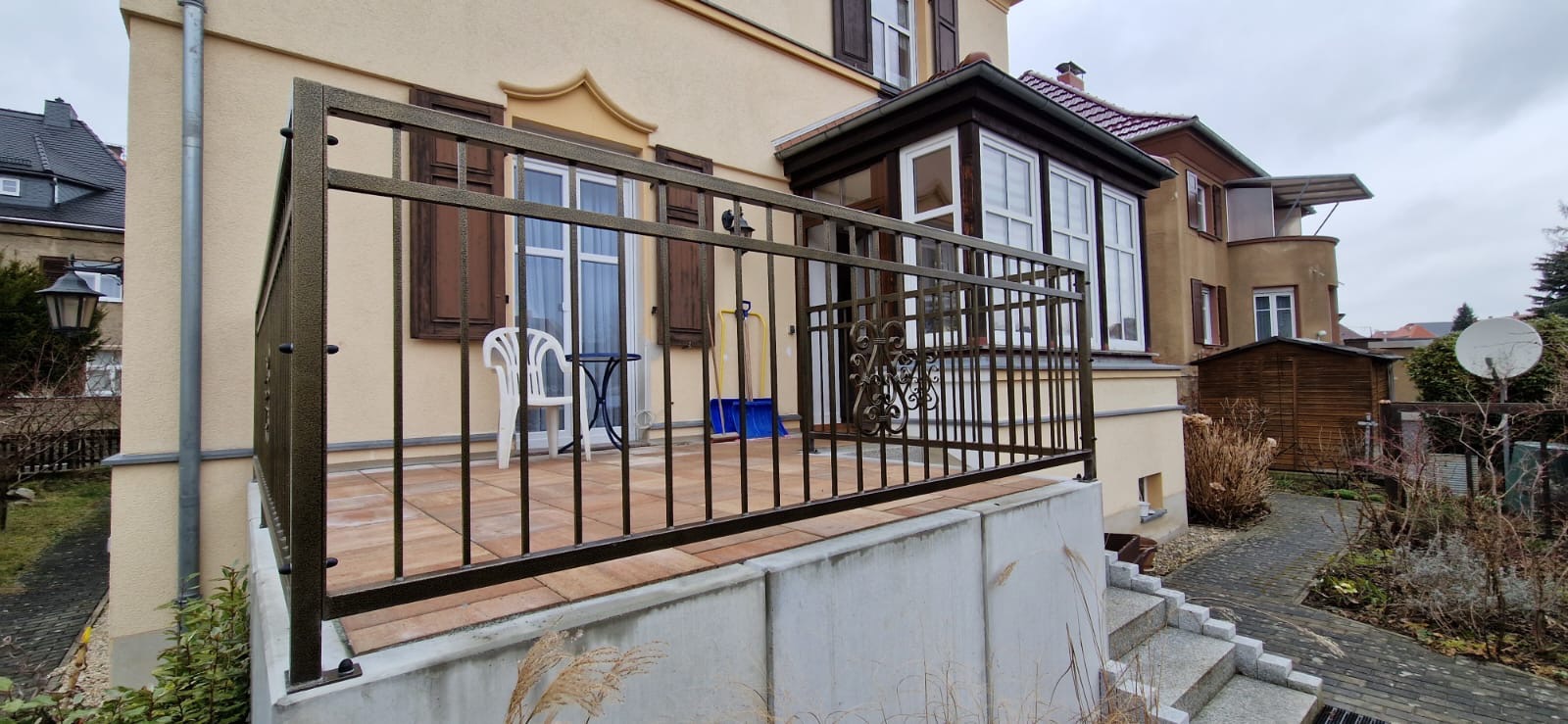 Metalowa balustrada balkonowa w kolorze brązu z dekoracyjnymi elementami, widok z dołu, w tle budynek mieszkalny z brązowymi okiennicami i białe krzesło z małym stolikiem na balkonie.