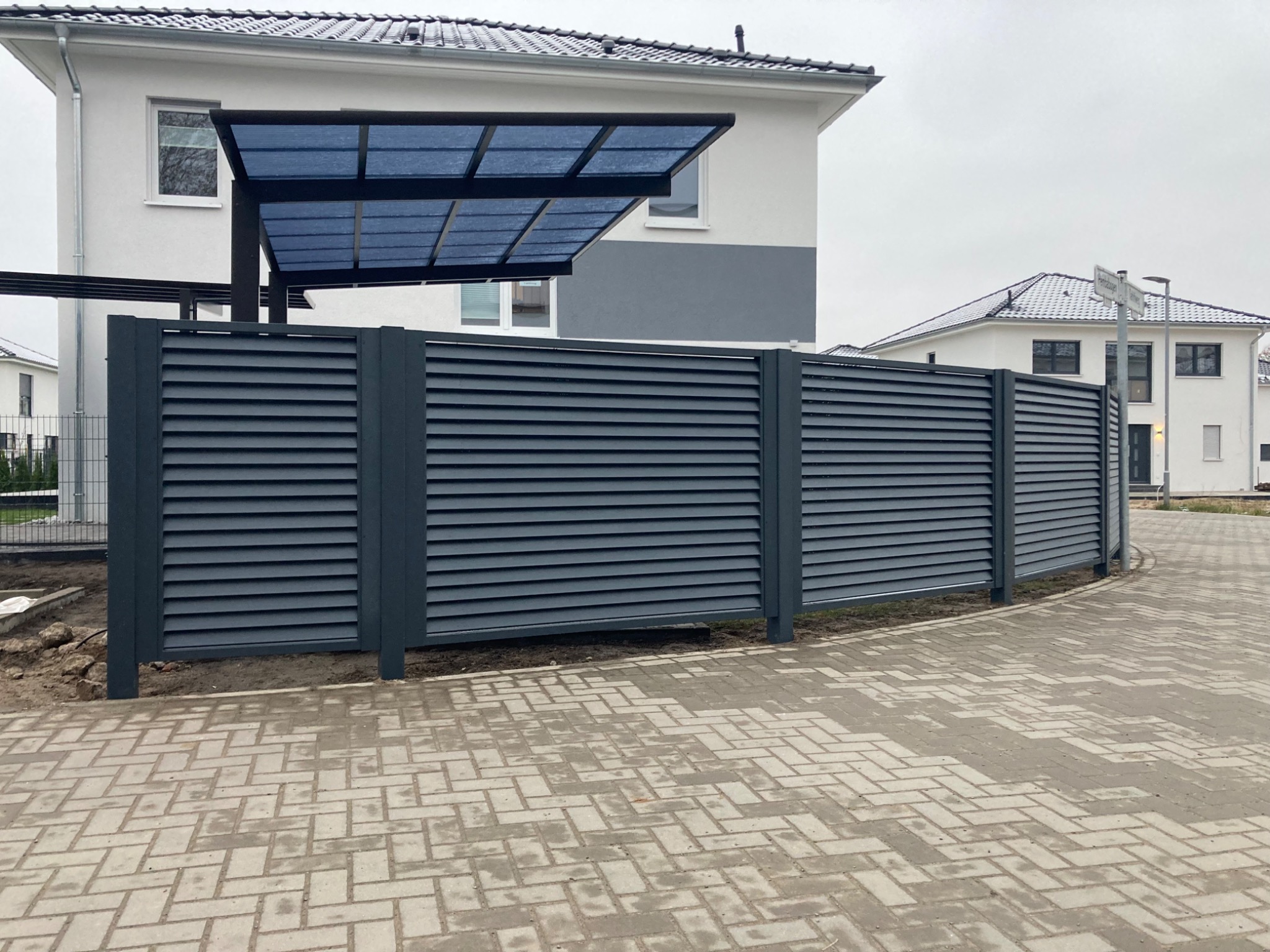 Nowoczesne, szare ogrodzenie z poziomych paneli na tle domu z wiatą garażową i kostki brukowej.