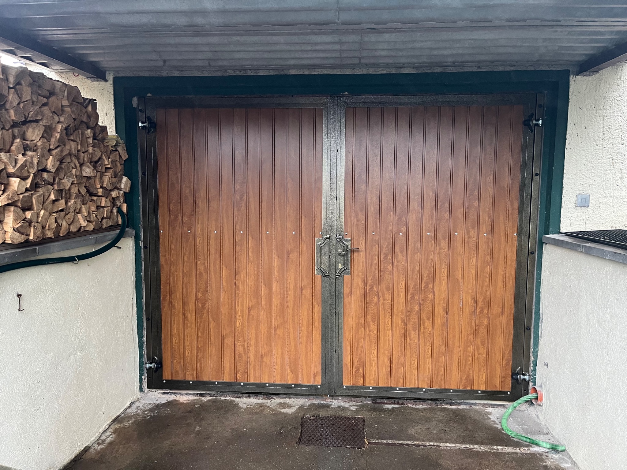 Dwuskrzydłowa brama z panelami imitującymi drewno, osadzona w metalowej ramie, z widocznym stosem drewna opałowego po lewej stronie.