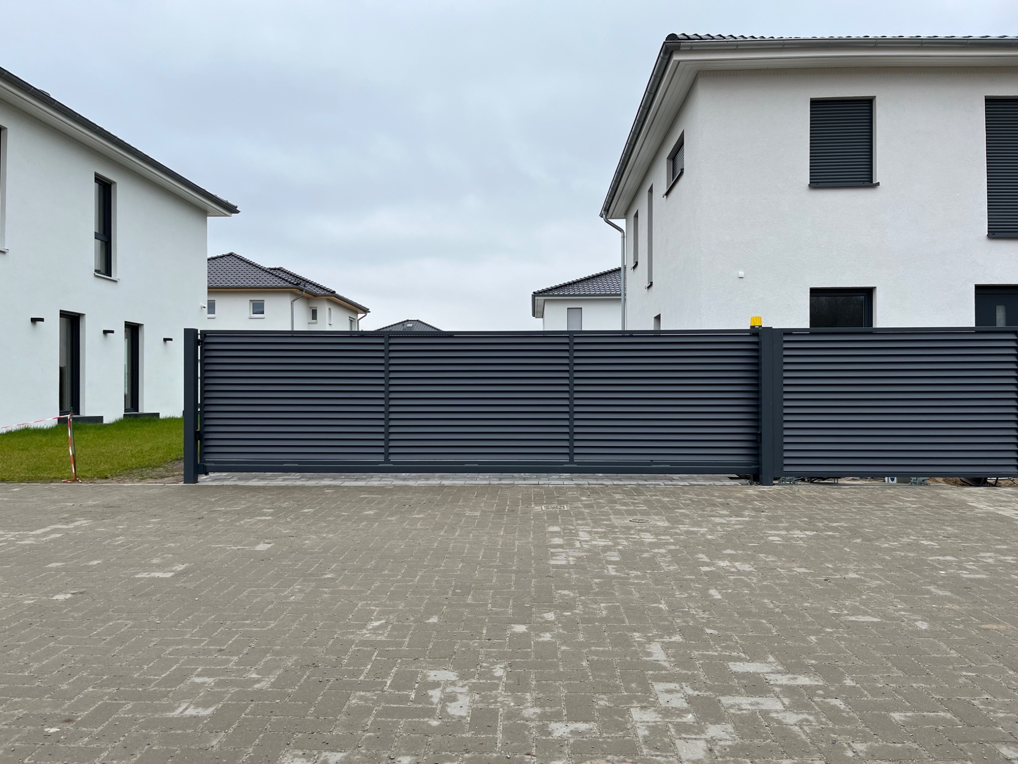 Nowoczesna, pozioma, przesuwna brama w kolorze antracytowym, z widocznymi białymi budynkami mieszkalnymi w tle, na tle pochmurnego nieba.