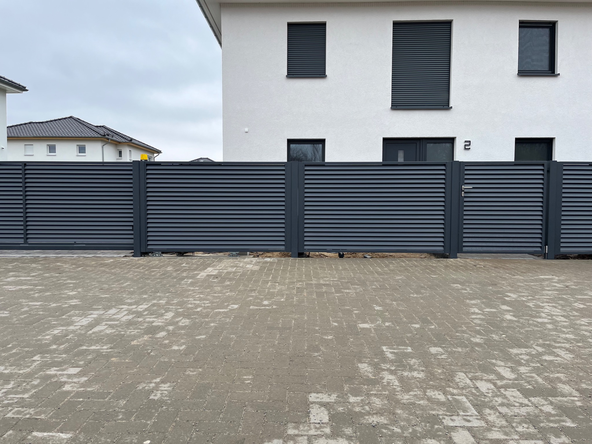 Nowoczesne, poziome przęsła ogrodzeniowe w kolorze grafitowym, zamykające posesję przed białym, dwupiętrowym domem z szarymi roletami w oknach, na pierwszym planie szara kostka brukowa.