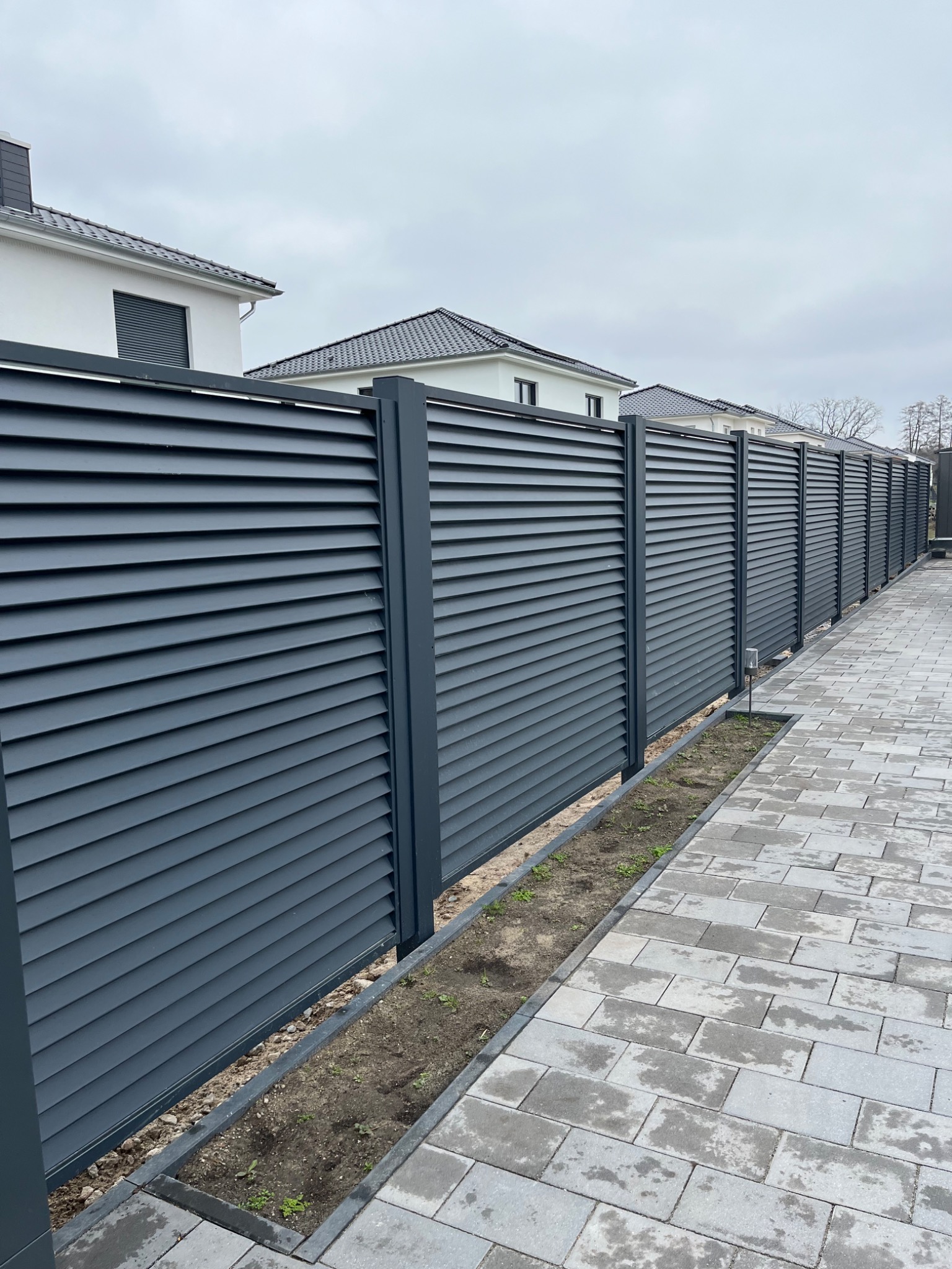 Długi, szary płot panelowy z poziomych listew, oddzielający posesję od chodnika z kostki brukowej, na tle białych domów z ciemnymi dachami w pochmurny dzień.