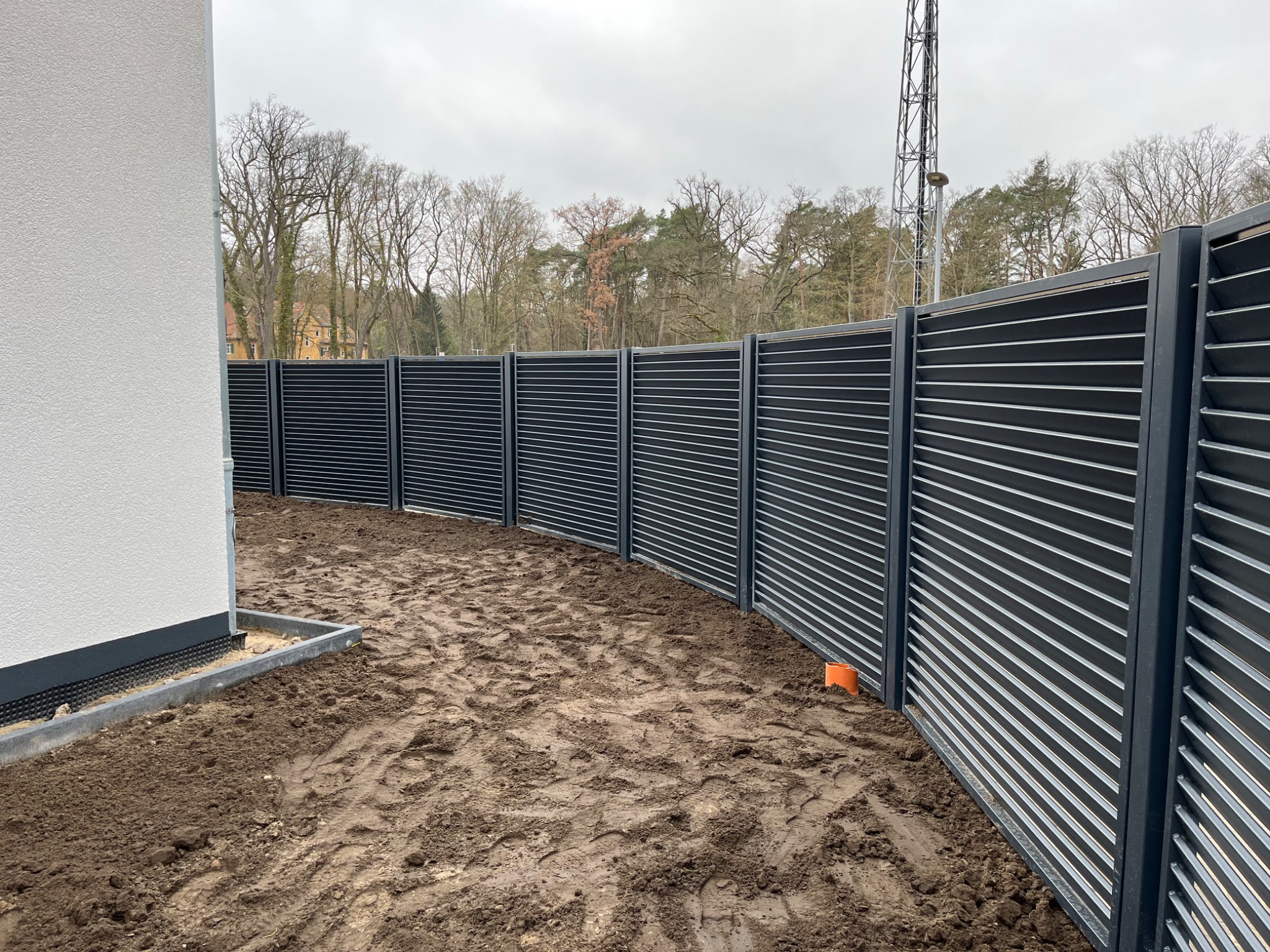 Nowoczesne, poziome przęsła ogrodzeniowe w kolorze grafitowym, tworzące łuk wokół posesji, z widoczną białą ścianą budynku i świeżo przekopaną ziemią.