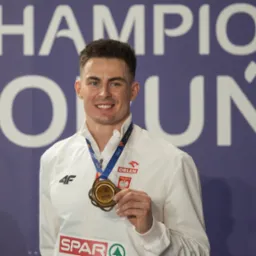 Uśmiechnięty sportowiec z medalem na tle napisu CHAMPION TORUŃ 2021, w białej bluzie z logo 4F i Orlen.