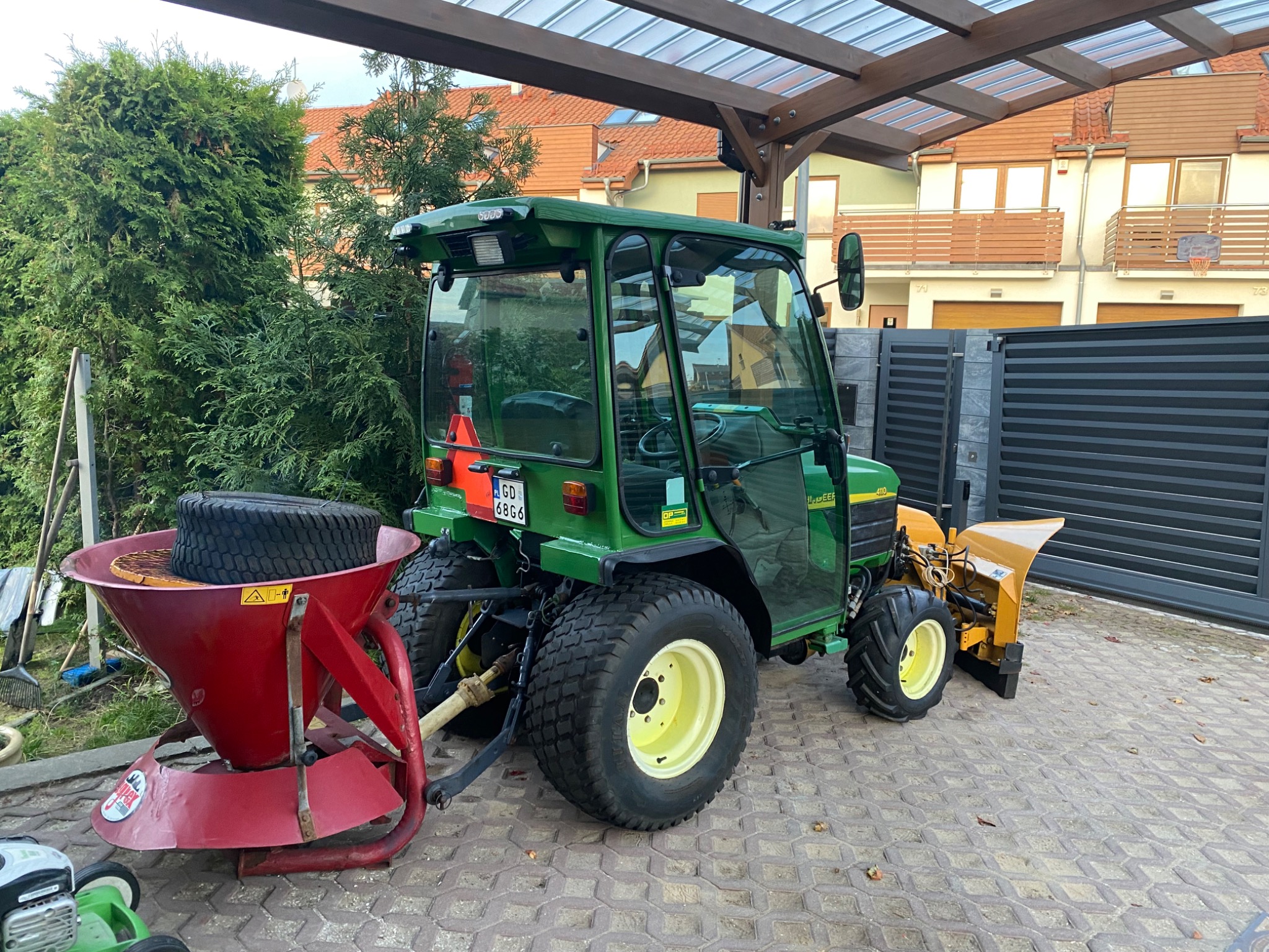 Zielony traktor z kabiną i odśnieżarką oraz rozsiewaczem nawozu lub soli stoi na kostce brukowej przed domem z szarą bramą.