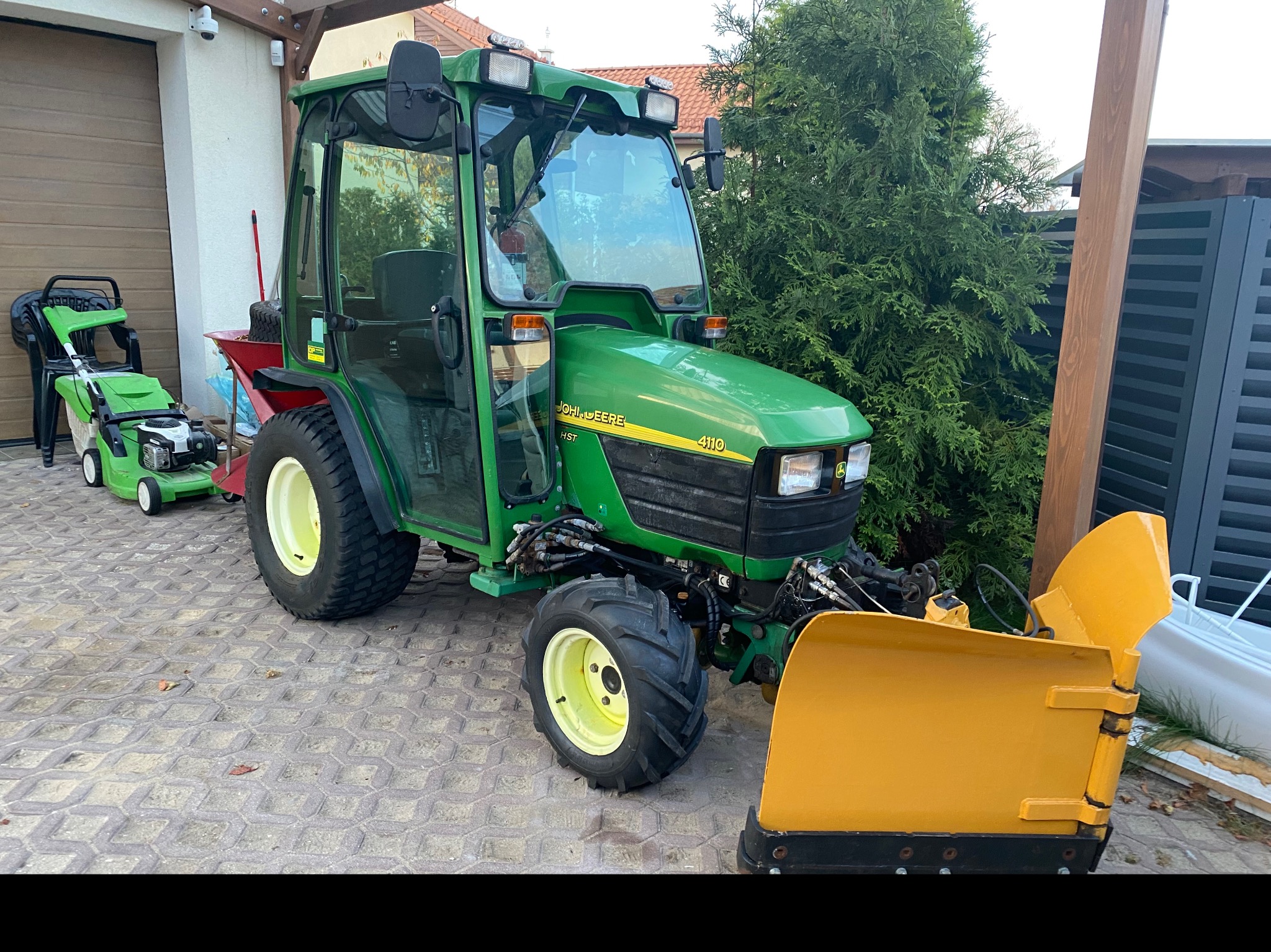 Zielony traktor John Deere 4110 z zamontowanym żółtym pługiem do śniegu, stojący na kostce brukowej, w tle garaż i zielony trawnik.
