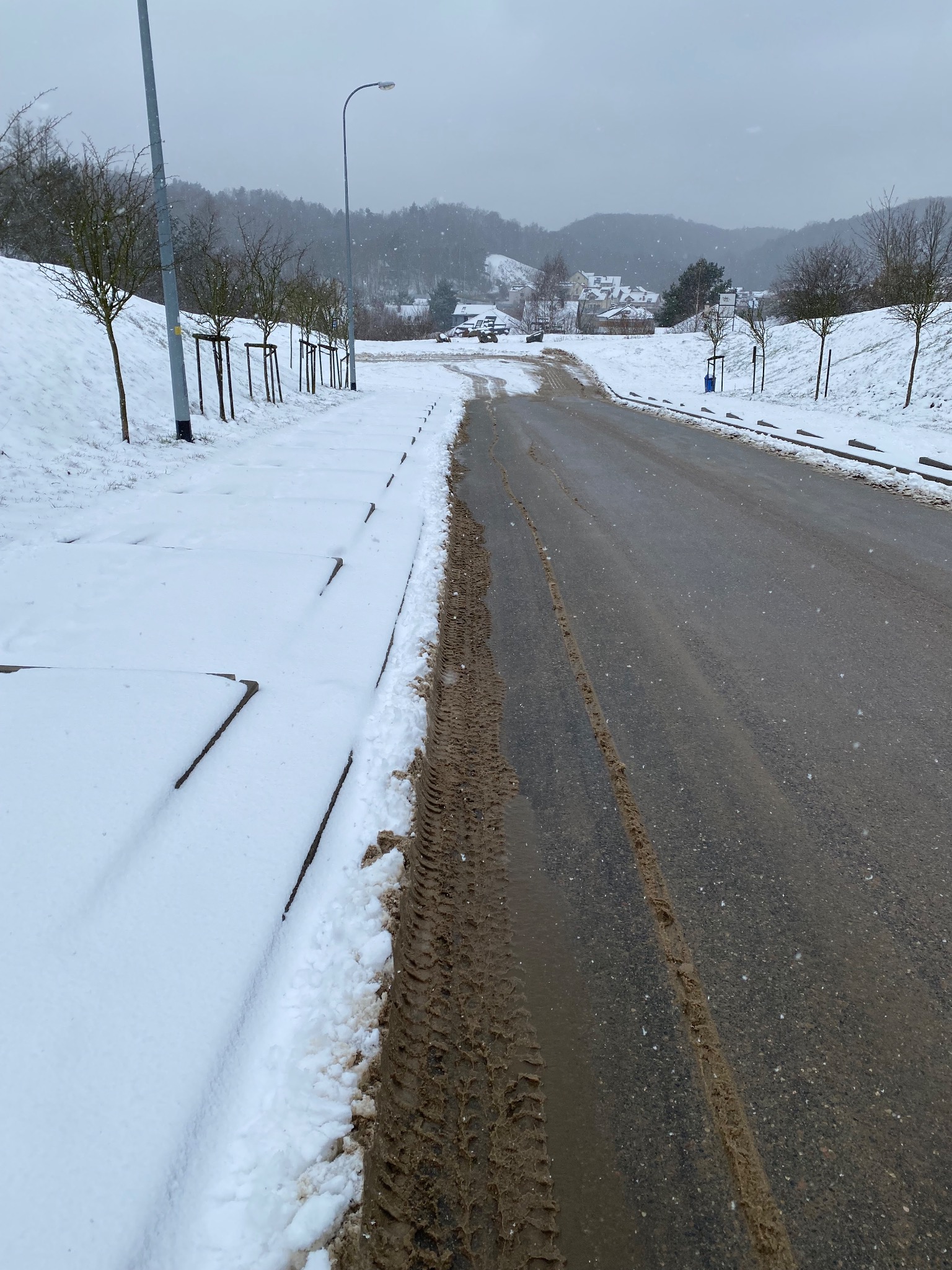 Ślady opon na mokrej, odśnieżonej drodze w zimowy dzień, z zasypanym śniegiem poboczem i widocznymi drzewami w tle.