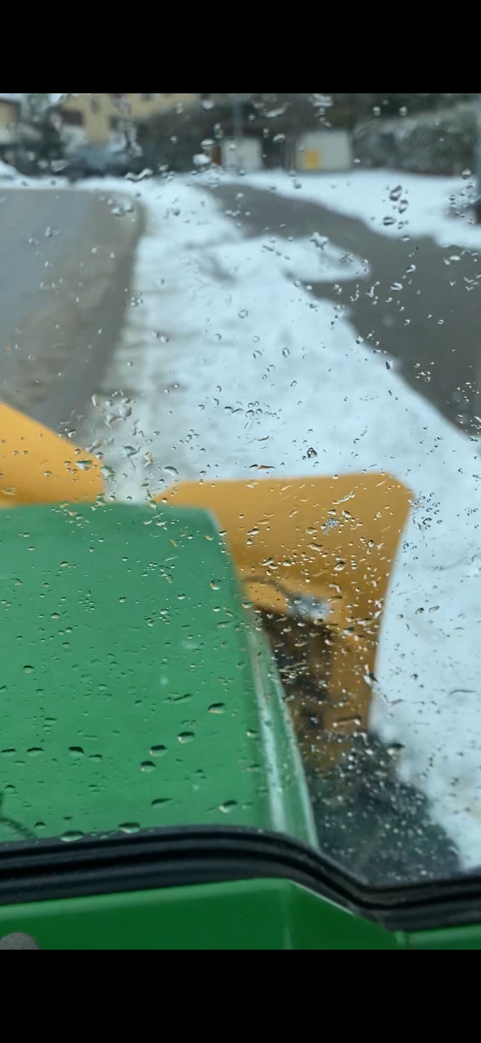 Widok z kabiny zielonego pojazdu na odśnieżaną drogę przez mokrą szybę, z żółtym pługiem w tle.