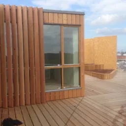 Zewnętrzny widok drewnianej konstrukcji budynku z drewnianą elewacją z pionowych desek, dużym oknem i drewnianym tarasem na dachu, z widokiem na zabudowania przemysłowe w tle.