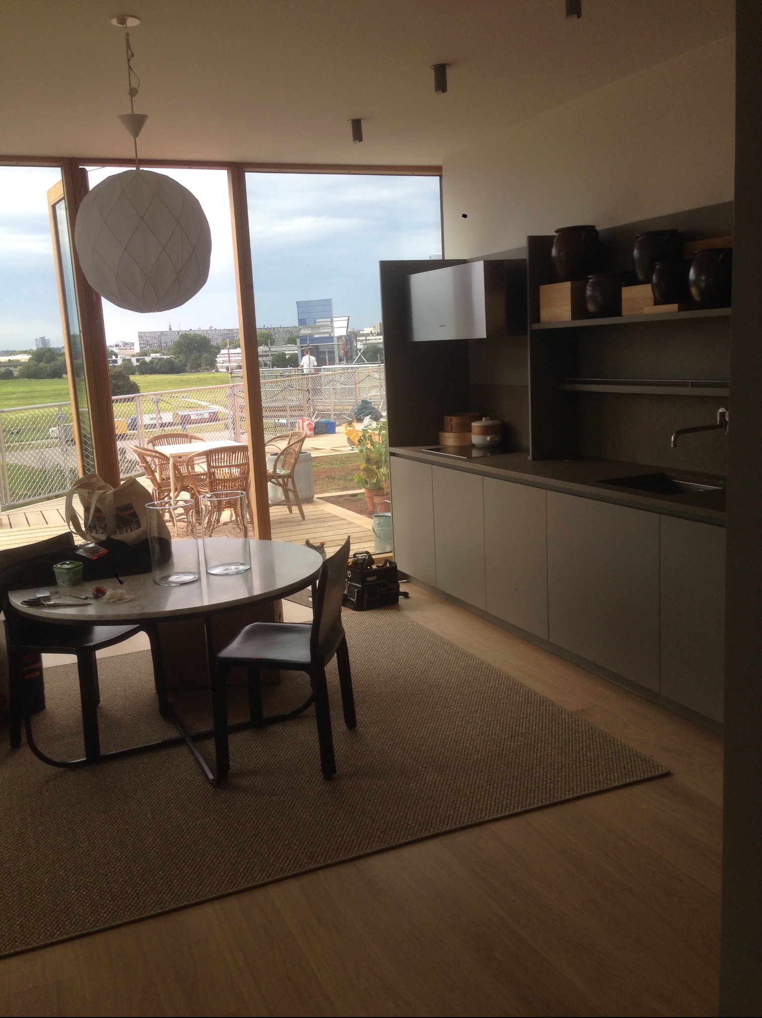 Jasne wnętrze domu z widokiem na zewnątrz przez duże okno, okrągły stół z krzesłami, minimalistyczna zabudowa kuchenna w odcieniach szarości, drewniana podłoga i dywan z naturalnego włókna.