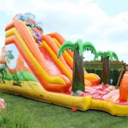 Dmuchana zjeżdżalnia z motywem dinozaurów i palm na trawniku, atrakcja dla dzieci na imprezie plenerowej.