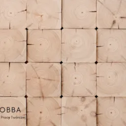 Drewniana ściana z kwadratowych bali z widocznymi słojami, logo 'HOBBA Dom Pracy Twórczej' w lewym dolnym rogu.