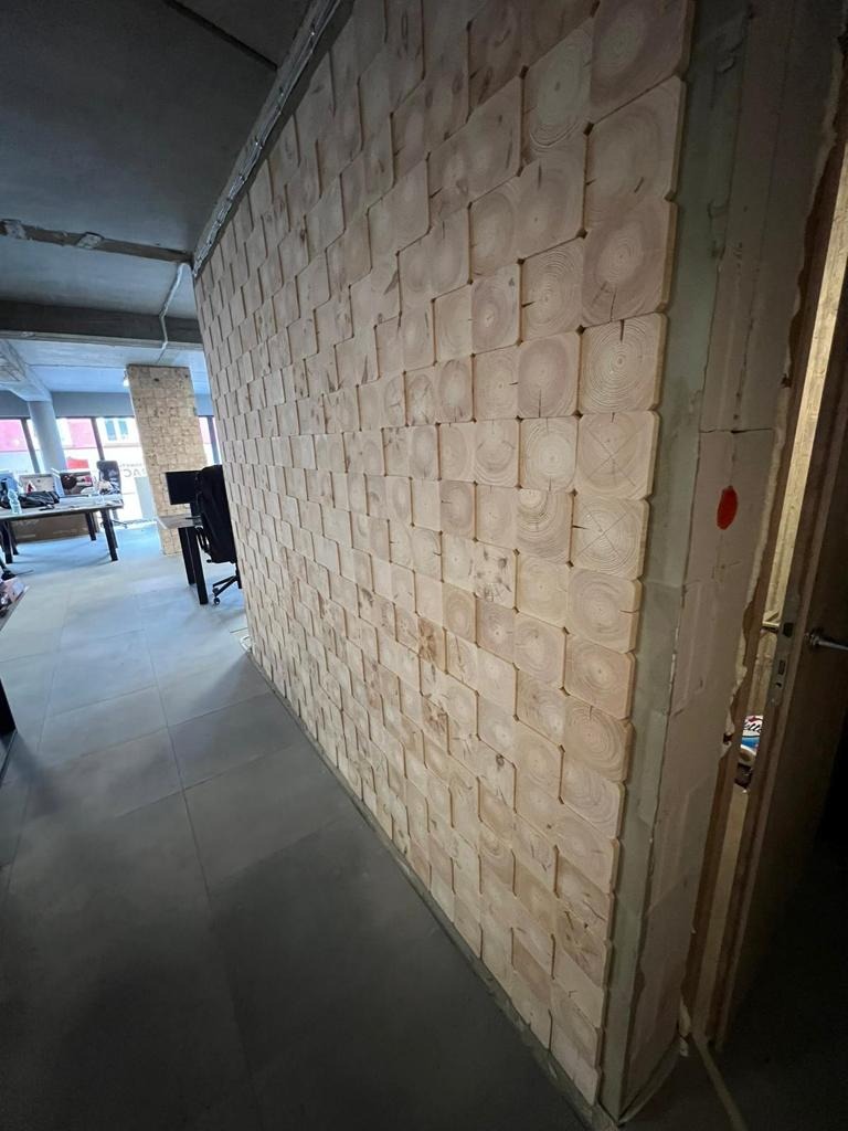 Ściana wykończona drewnianymi kostkami z widocznymi słojami, surowy betonowy sufit, szara podłoga, widok na biuro w tle.