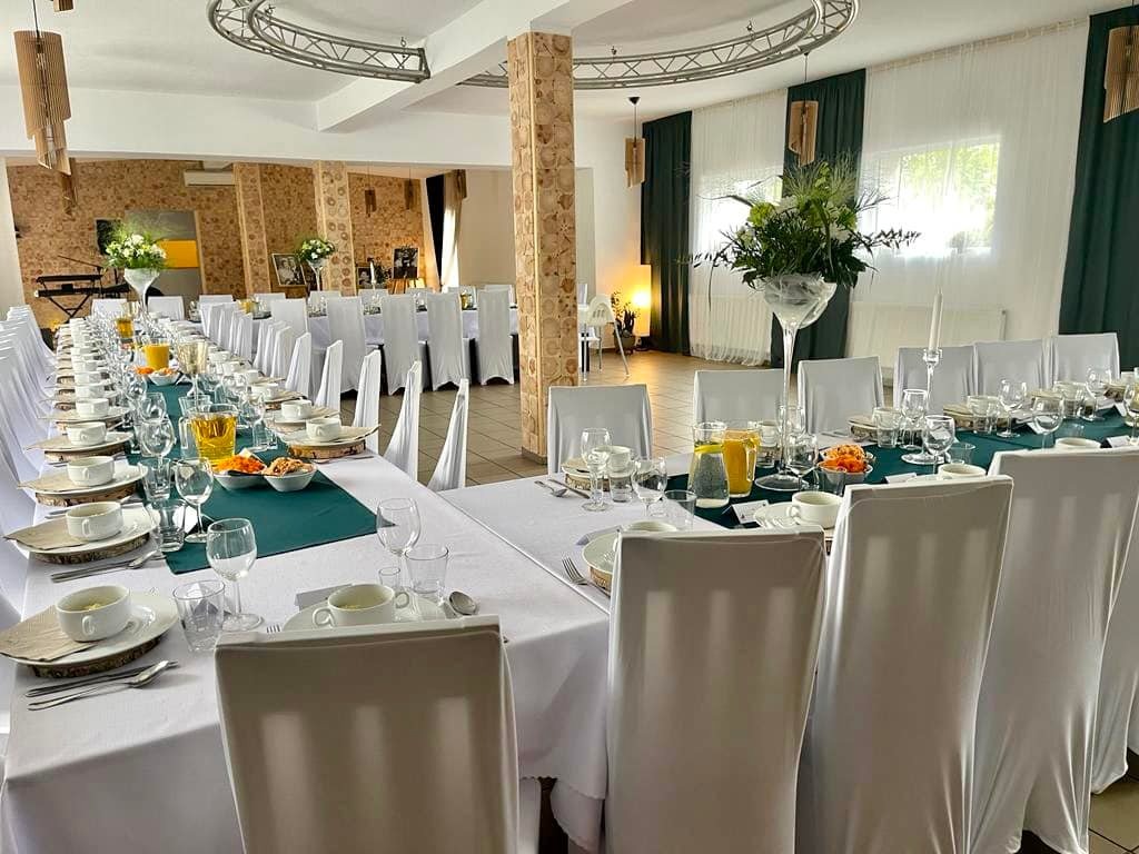 Uroczysta sala bankietowa z długim stołem nakrytym białym obrusem, zielonym bieżnikiem, zastawą, szklankami i dzbankami z sokiem, udekorowana kwiatami, z krzesłami w białych pokrowcach, kolumna...