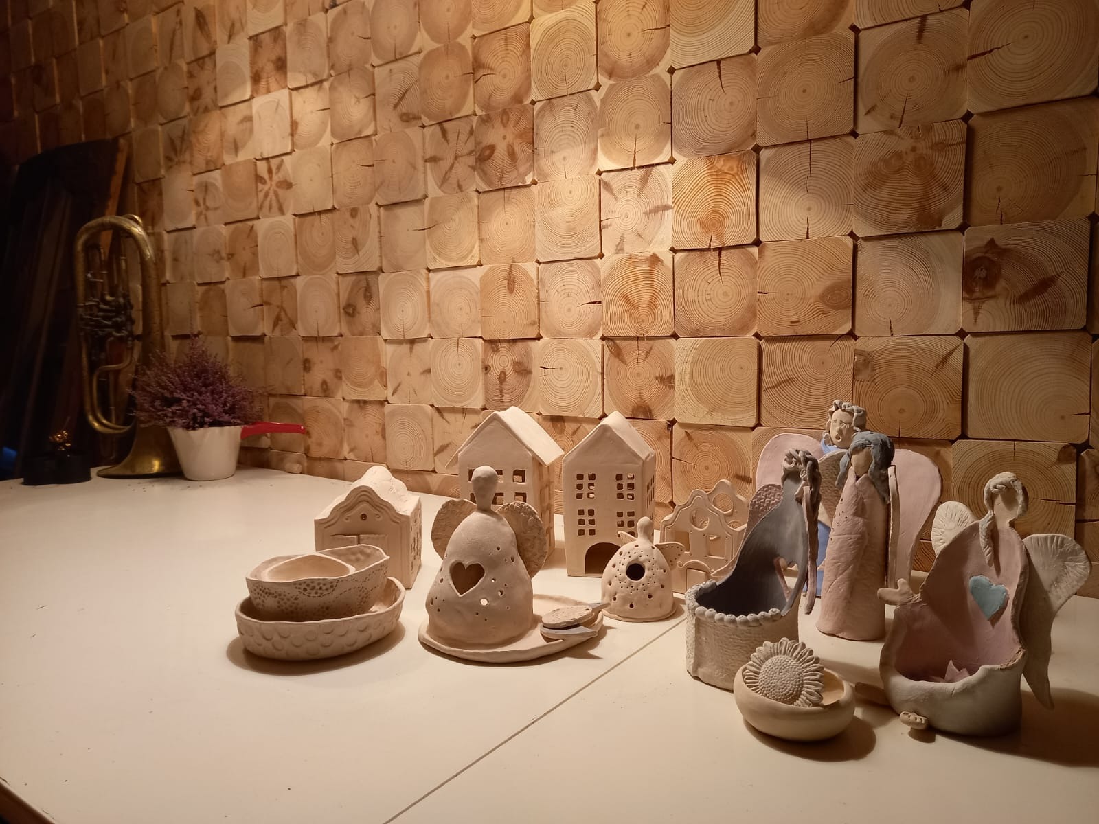 Ściana wykończona drewnianymi klockami, przed nią ręcznie robione ceramiczne figurki aniołów, domków i miseczek, obok wazon z wrzosem i stara trąbka.