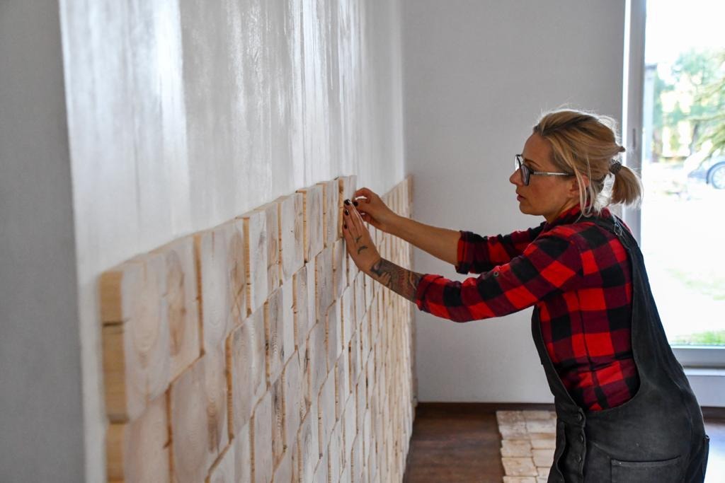 Kobieta w okularach i czerwonej koszuli w kratę układa dekoracyjne panele ścienne z drewnianych klocków na białej ścianie, widoczny fragment podłogi z tego samego materiału.