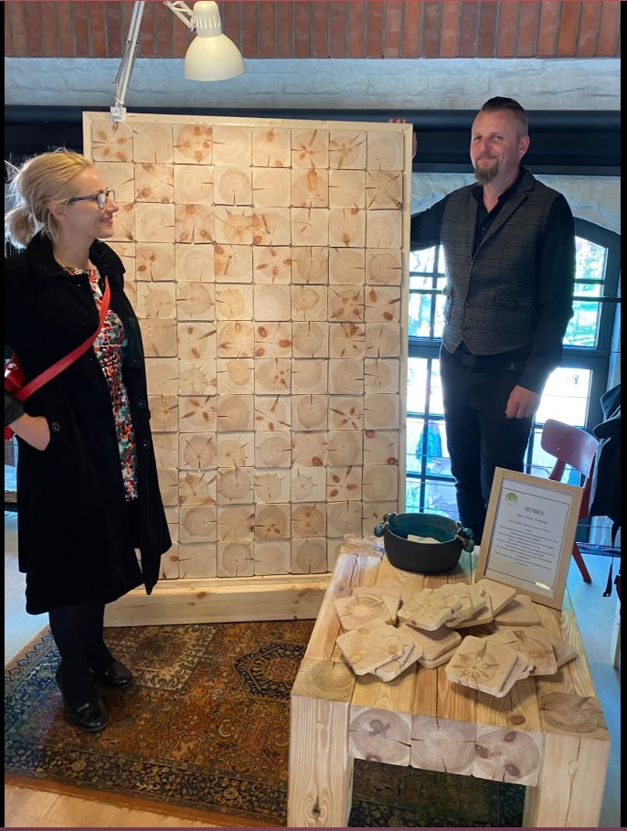 Panel ścienny z drewnianych kostek z widocznymi słojami drewna, prezentowany przez kobietę w okularach i mężczyznę w kamizelce, ustawiony na tle ceglanej ściany.