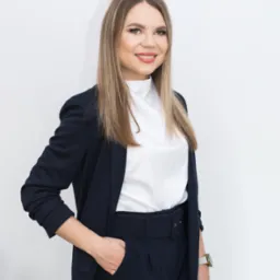 Uśmiechnięta kobieta w granatowym garniturze i białej bluzce, stojąca na tle białej ściany, z ręką w kieszeni spodni i zegarkiem na nadgarstku.