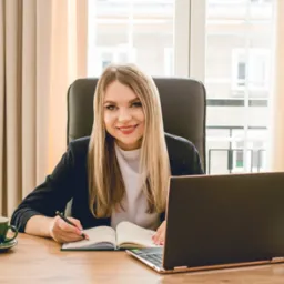 Uśmiechnięta kobieta w biurze, pisząca w notatniku obok laptopa, z filiżanką kawy i kodeksami na biurku, w tle okno z widokiem na budynek.