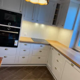 Jasna kuchnia z drewnianym blatem, fronty szafek z frezowanymi ramkami i mosiężnymi uchwytami, zabudowany piekarnik i płyta indukcyjna, dekoracyjna lampa z abażurem z rattanu.