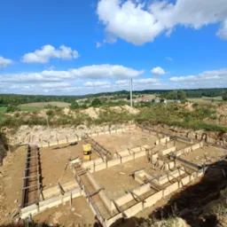 Widok z góry na przygotowany fundament pod budynek z drewnianą konstrukcją szalunkową i zbrojeniem, na tle błękitnego nieba z białymi chmurami i odległego krajobrazu.