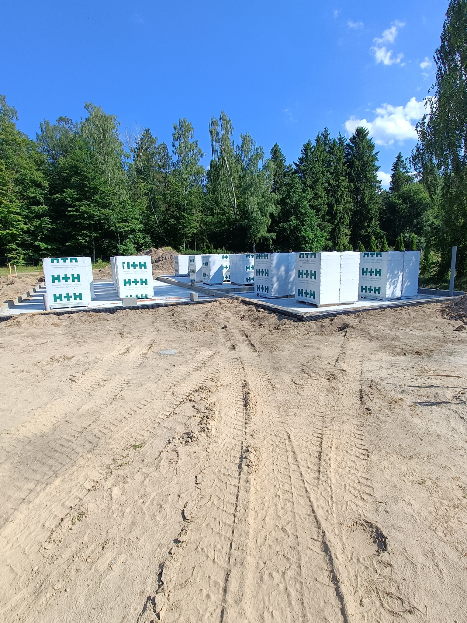 Przygotowany fundament pod budowę domu z bloczków H+H, widok na teren budowy z wyraźnymi śladami opon w piasku, w tle las.