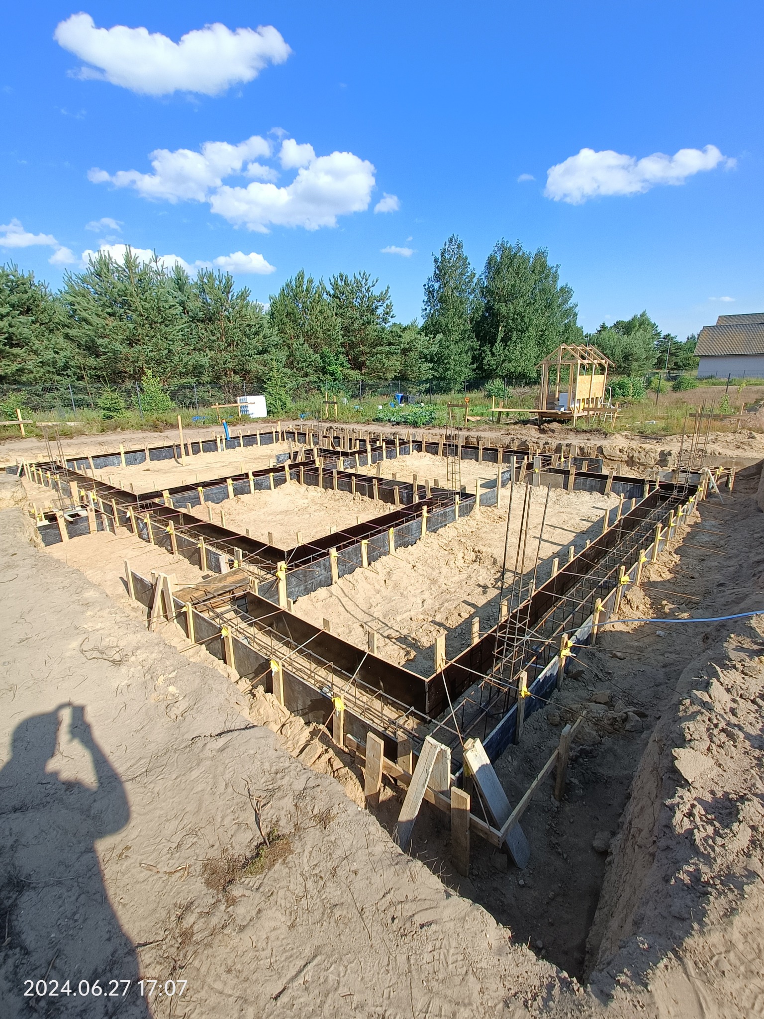 Przygotowanie fundamentów pod budowę domu z widoczną konstrukcją szalunkową, zbrojeniami i wykopem, na tle drzew i błękitnego nieba z chmurami.