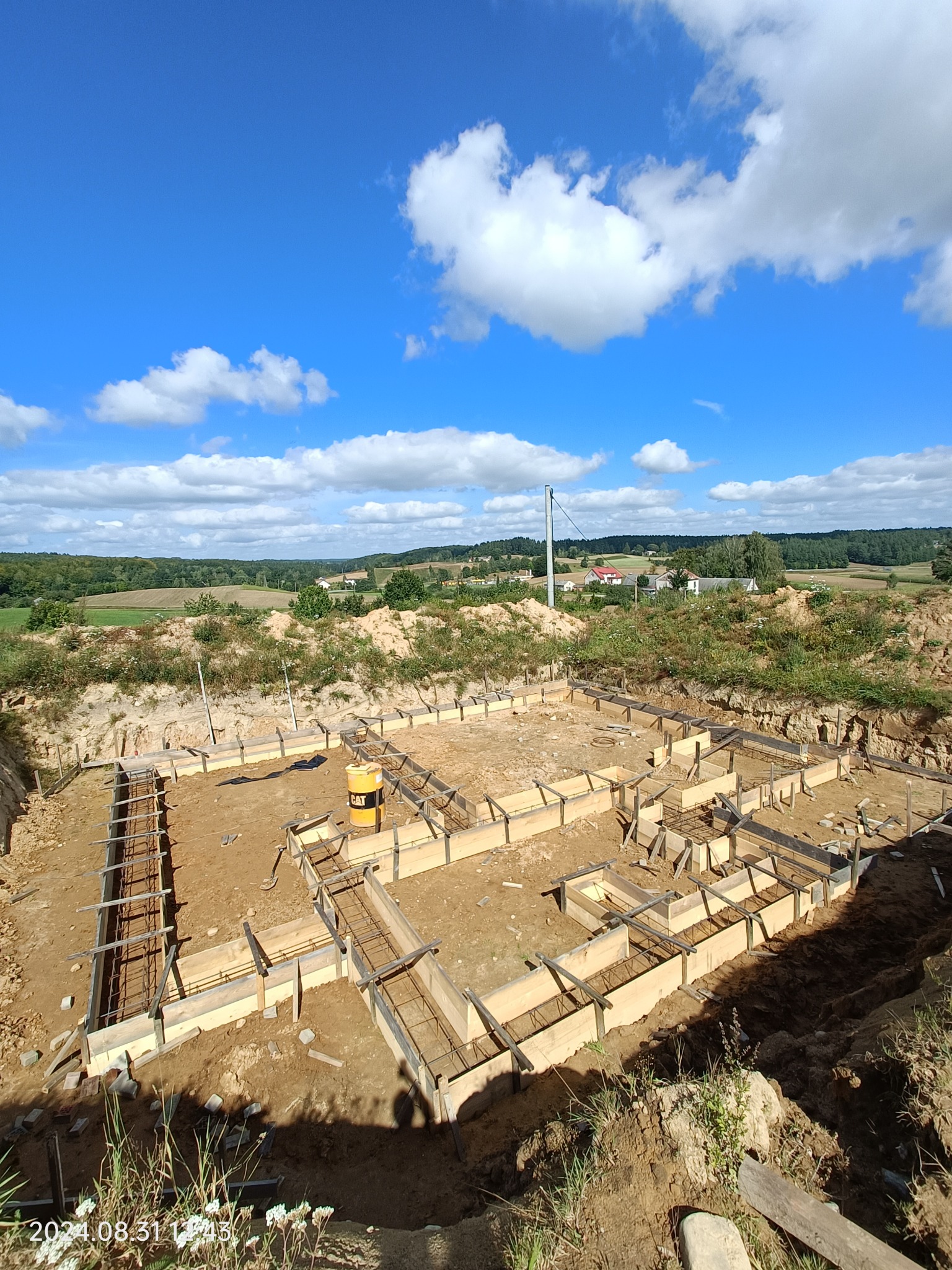 Widok z góry na przygotowany fundament pod budynek z drewnianą konstrukcją szalunkową i zbrojeniem, na tle błękitnego nieba z białymi chmurami i odległego krajobrazu.