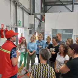 Grupa osób słucha instruktora w czerwonej kamizelce i czapce podczas szkolenia z obsługi hydrantu wewnętrznego w hali przemysłowej.