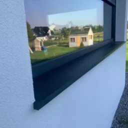 Czarny parapet zewnętrzny z konglomeratu zamontowany w oknie nowoczesnego domu, z widokiem na ogród i domek narzędziowy odbijającym się w szybie.