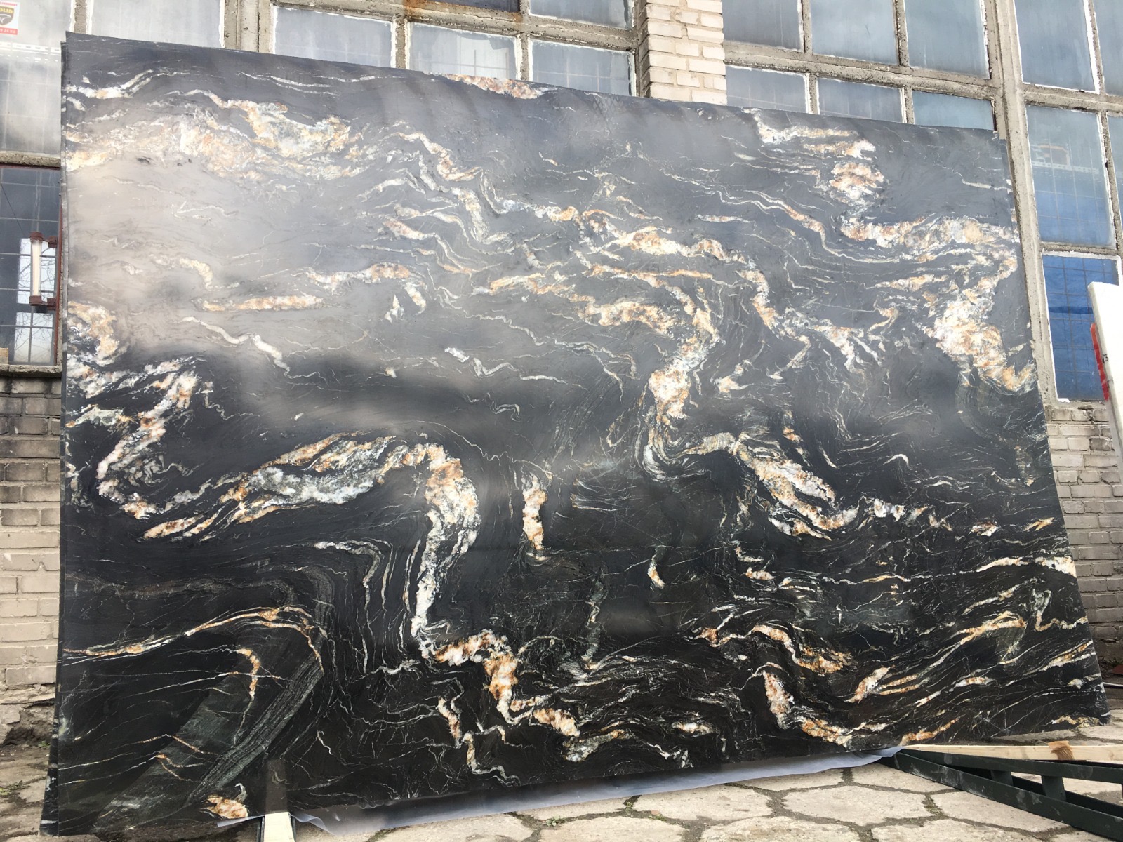 Duża płyta czarnego kamienia z wyraźnymi, falistymi żyłami w kolorach bieli i złota, oparta o ceglany budynek z oknami.