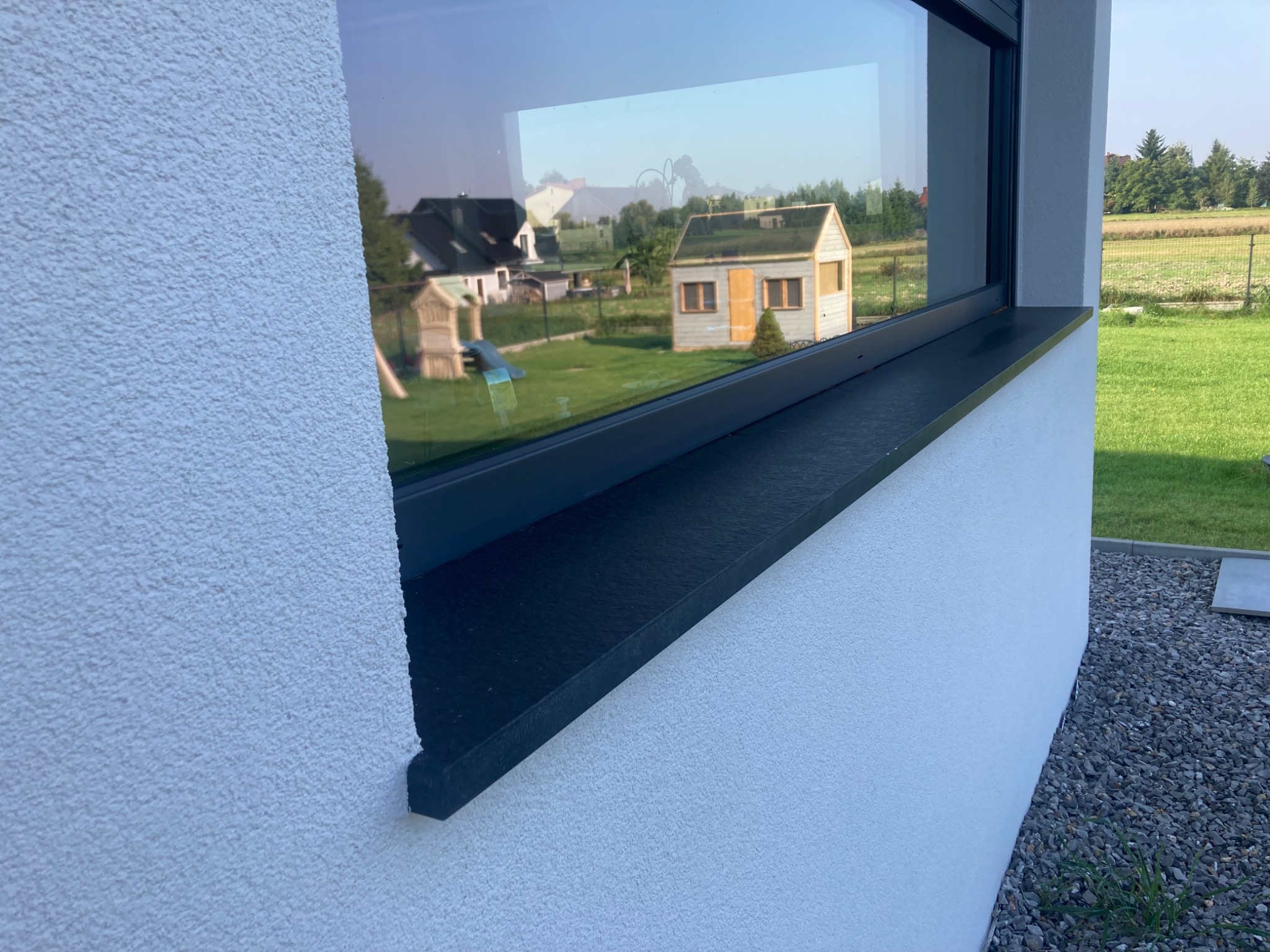 Czarny parapet zewnętrzny z konglomeratu zamontowany w oknie nowoczesnego domu, z widokiem na ogród i domek narzędziowy odbijającym się w szybie.