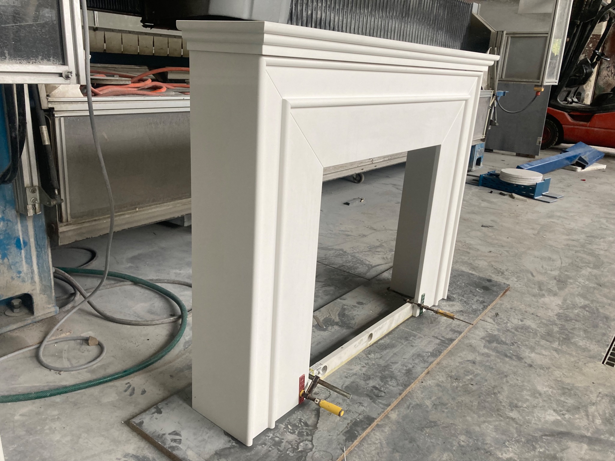 Biały, klasyczny portal kominkowy z konglomeratu, w trakcie montażu, zabezpieczony ściskami stolarskimi, na tle hali produkcyjnej z widocznym wózkiem widłowym.