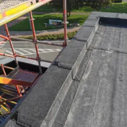 Widok z góry na płaski dach pokryty papą, obok rusztowanie z pomarańczowych rur, w tle zieleń trawników i drzewa.