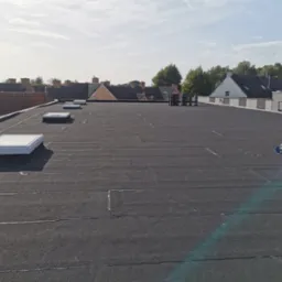 Czarny, płaski dach pokryty papą termozgrzewalną z widocznymi świetlikami dachowymi oraz rolkami papy w tle, na tle zabudowań mieszkalnych i błękitnego nieba.