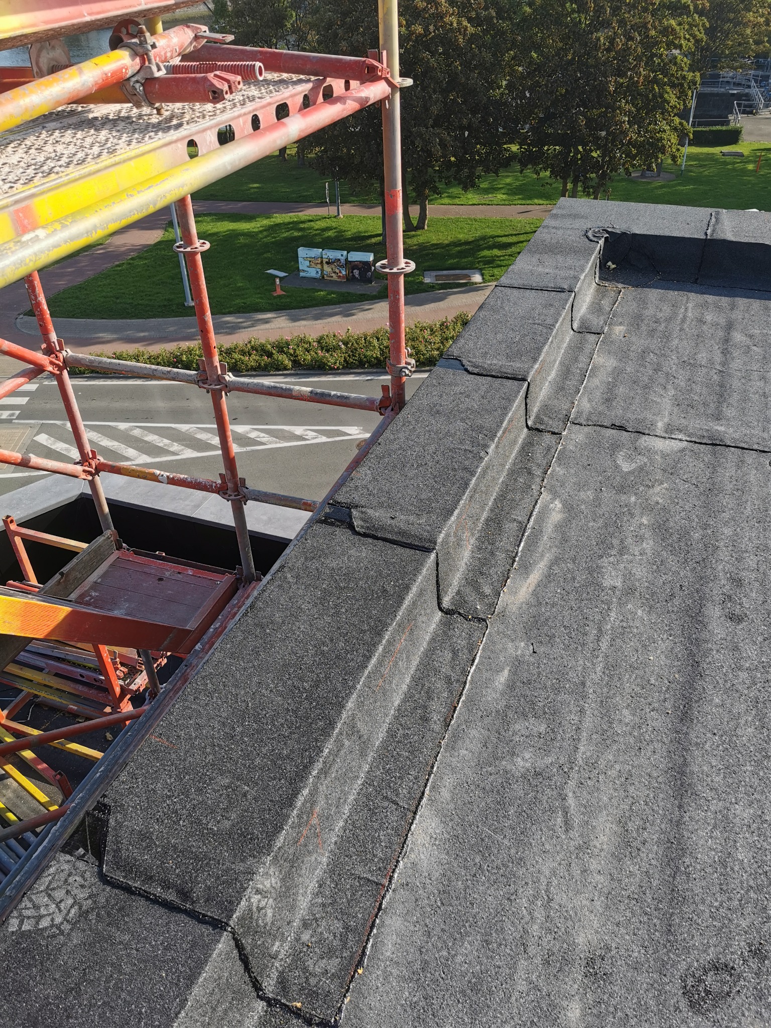 Widok z góry na płaski dach pokryty papą, obok rusztowanie z pomarańczowych rur, w tle zieleń trawników i drzewa.