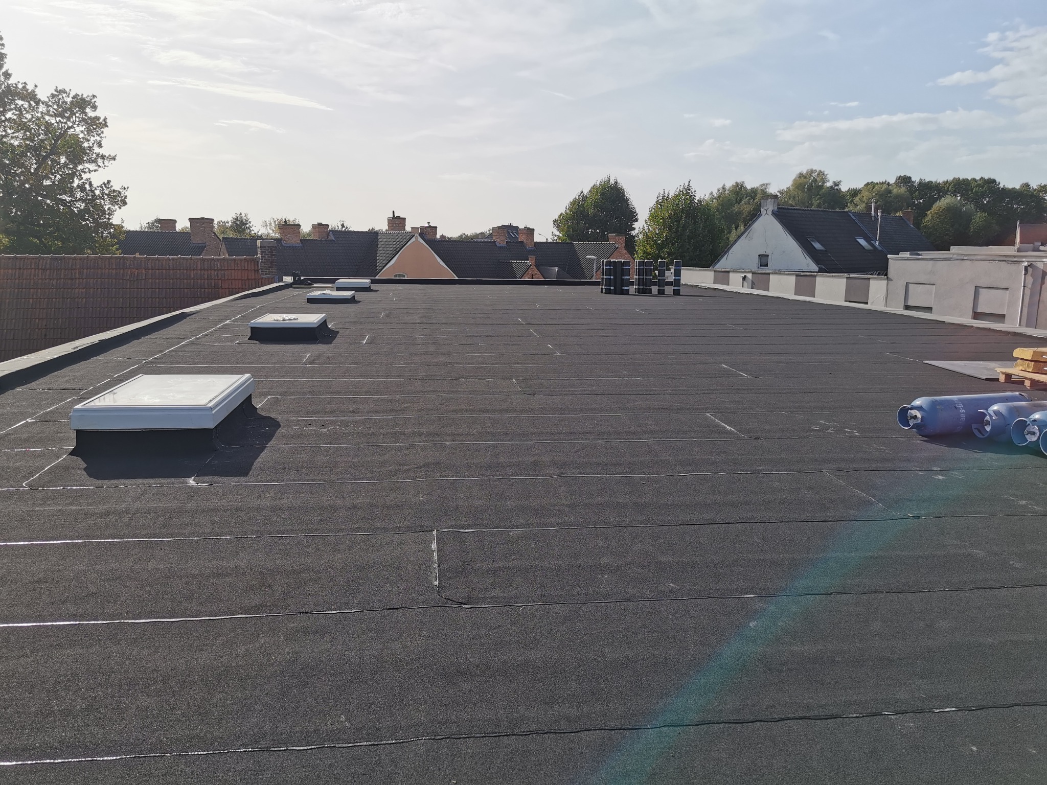 Czarny, płaski dach pokryty papą termozgrzewalną z widocznymi świetlikami dachowymi oraz rolkami papy w tle, na tle zabudowań mieszkalnych i błękitnego nieba.