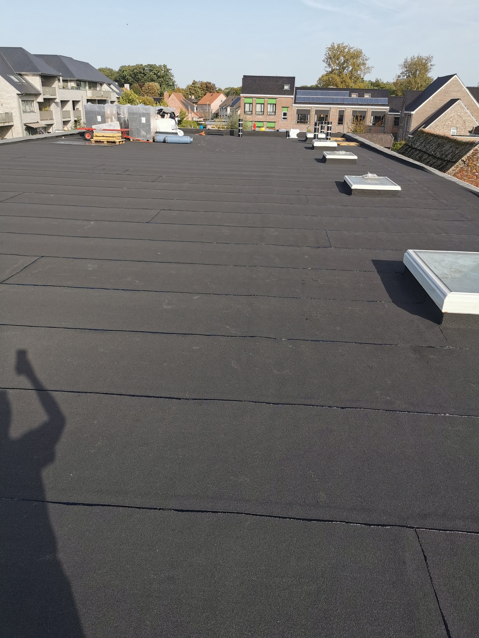 Nowe, czarne pokrycie dachowe z papy bitumicznej na płaskim dachu, widoczne świetliki dachowe i cień fotografa w lewym dolnym rogu.