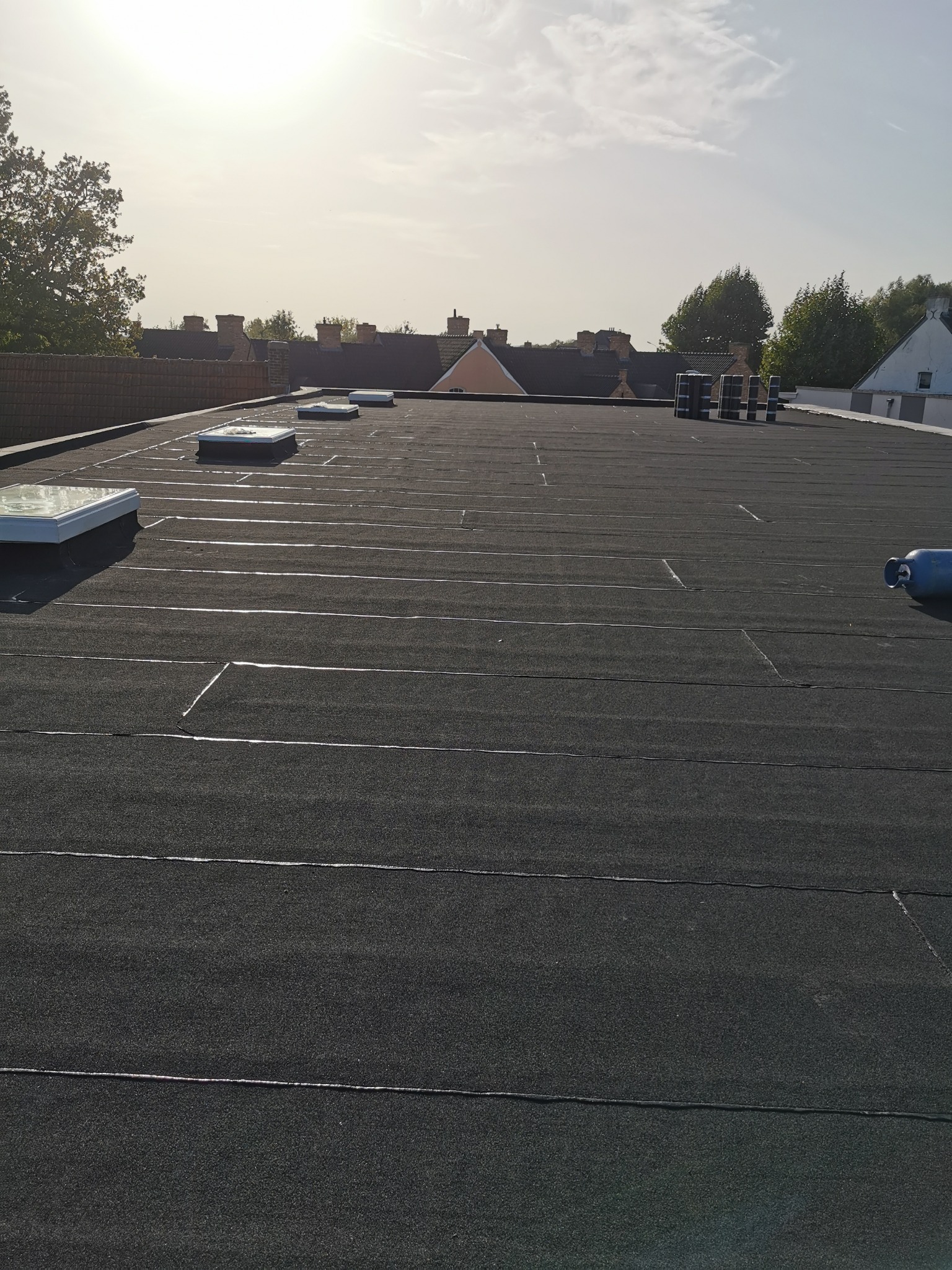 Widok z góry na płaski dach pokryty czarną papą z widocznymi łączeniami, świetlikami i butlą gazową w rogu; w tle inne budynki i drzewa pod jasnym niebem.