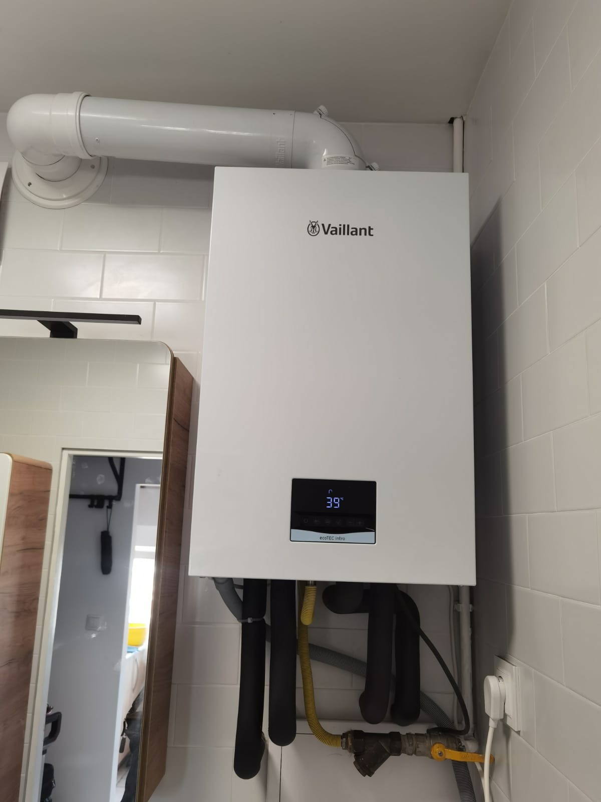 Biały, wiszący piec gazowy marki Vaillant, model ecoTEC intro, zamontowany na ścianie z białych płytek, z widocznymi rurami instalacji i fragmentem lustra po lewej stronie.