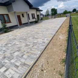 Nowoczesny podjazd z kostki brukowej w odcieniach szarości i beżu prowadzący do domu z beżową elewacją i ciemną stolarką okienną, widok na posesję z metalowym ogrodzeniem.